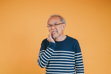 Fototapeta premium Dental pain. Portrait senior old man sad hand touching cheek suffering from toothache studio shot isolated on yellow background, Asian unhappy elder man problems with teeth pain, dental healthcare