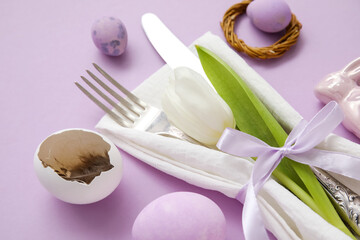 Table serving with Easter eggs and tulip flower on lilac background, closeup