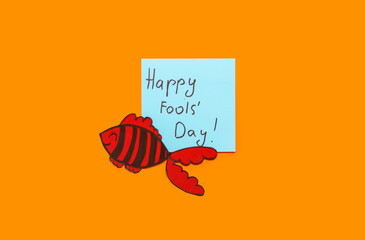 Sticky note with text HAPPY FOOL'S DAY and paper fish on color background