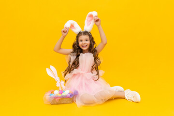 Obraz na płótnie Canvas Festive Easter. A curly-haired girl with rabbit ears in a pink dress is enjoying the holiday. A child with a basket of colorful eggs on a yellow isolated background.