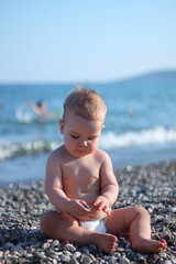 Fototapeta na wymiar A cute chubby baby boy blonde in a diaper sits on a pebble against the background of the sea and looks at a pebble in his hand.