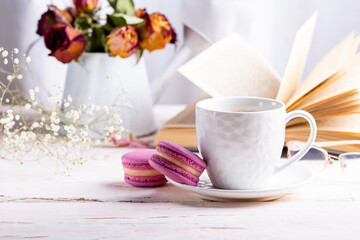 Fototapeta na wymiar French macaroon dessert. Bright pink macaroons and a white cup of coffee on a white wooden table against the backdrop of an open book and a winter bouquet of dried flowers