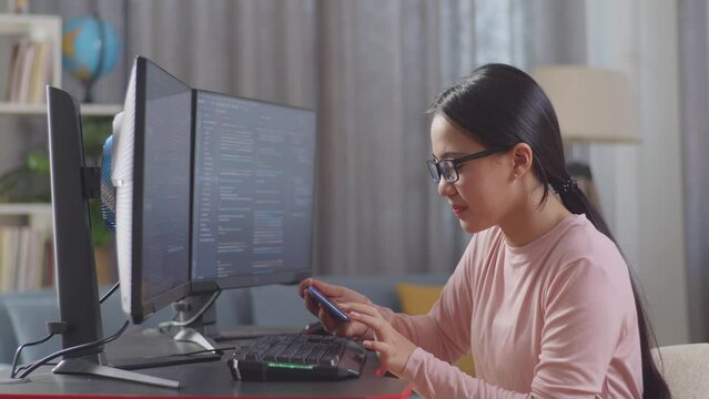 Asian Teen Girl Programmer Using Smartphone While Creating Software Engineer Developing App, Program, Video Game On Destop Computer At Home
