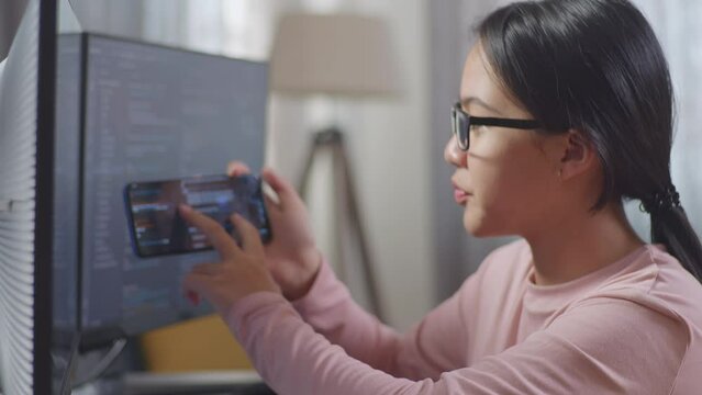 Close Up Of Asian Teen Girl Programmer Looking At Smartphone Then Celebrating The Success While Creating Software Engineer Developing App, Program, Video Game On Desktop Computer At Home
