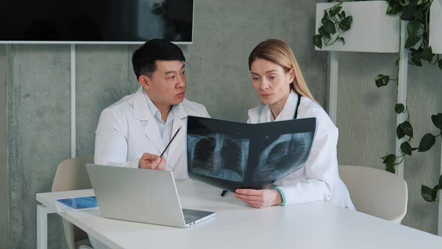 Two Diverse Doctors Talk and Use Laptop, Analysing Patient X-ray, Talking about Results Analysis Health Care Clinic, Asian Chief Surgeon and Radiologist Discuss Consult about Image of Patient's Lungs.