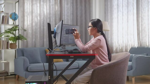 Asian Teen Girl Programmer Using Smartphone While Creating Software Engineer Developing App, Program, Video Game On Destop Computer At Home
