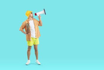 Young man wearing summer shirt, shorts, sun hat and sunglasses standing on blank turquoise copyspace studio background, holding pineapple and megaphone and making announcement about great holiday sale