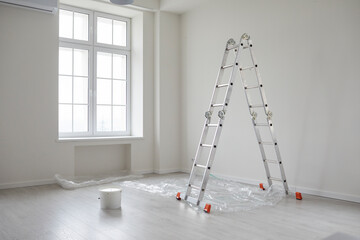 Modern house or apartment after home renovations. Empty, clean living room interior with white freshly painted walls, ladder, paint can, protective plastic on the floor, big window, and no people