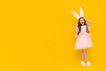 Festive Easter. A teenage girl in a fluffy dress and with rabbit ears points to your advertisement on a yellow isolated background. A child in a rabbit costume stands tall. Copy space.