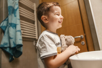 A boy is smiling at the mirror while standing in the bathroom with his bunny toy in the morning.