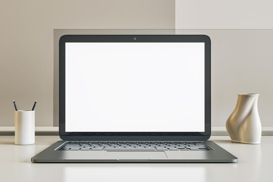 Front view on blank white modern laptop monitor screen with space for web design or web page on glossy surface table among stand for pens and vase on light wall background. 3D rendering, mockup