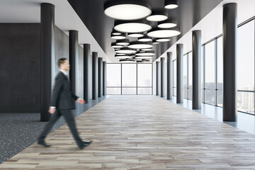 Fototapeta premium Side view on blurred walking businessman on wooden floor in spacious empty business center corridor with stylish lamps on dark top, black pillars in front panoramic windows with city view background
