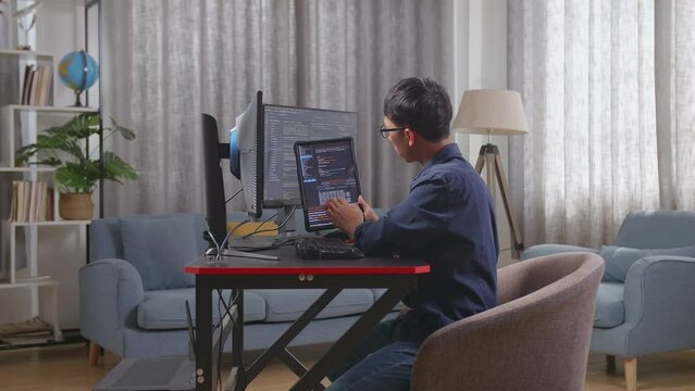 Asian Boy Programmer Looking At Database On Tablet While Creating Software Engineer Developing App, Program, Video Game On Desktop Computer At Home

