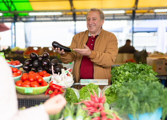 Smiling mature male seller passing eggplants through counter to buyer at local market