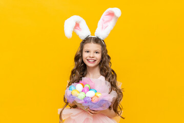 Obraz na płótnie Canvas Festive Easter for children. A beautiful teenage girl with curly hair holds a basket of colored colored eggs for the holiday. A child in a rabbit costume with long ears on a yellow isolated background