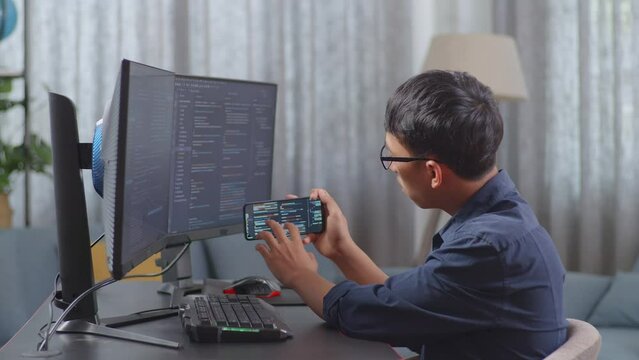 Asian Boy Programmer Using Smartphone While Creating Software Engineer Developing App, Program, Video Game On Destop Computer At Home
