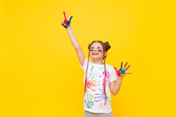 A beautiful little girl with stained hands in paint points her fingers in different directions. A child in paint on a yellow background.