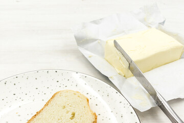 Fresh piece of butter on the packaging paper, knife and a plate of bread.