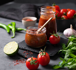 Hot sauce adjika. Homemade appetizer with peppers and tomatoes in a jar on a dark background with vegetables and herbs. Rustic style.