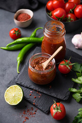 Hot sauce adjika. Homemade appetizer with peppers and tomatoes in a jar on a dark background with vegetables and herbs. Rustic style.