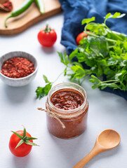 Hot sauce adjika. Homemade appetizer with pepper and tomatoes in a jar on light background with fresh herb and vegetables.