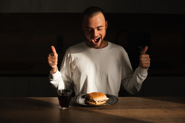 Attractive amazed surprised man show thumbs up gesture and sitting near table with plate with burger. Fast food. Male wear white long sleeves sitting at house in kitchen and look at burger, open mouth
