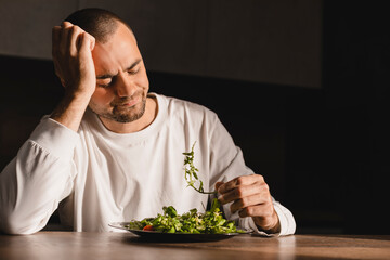 Man sitting at table, looking sad and bored with diet not wanting to eat salad. Modern kitchen interior. Healthy nutrition, dieting, eating disorder. Man look at salad unhappy.