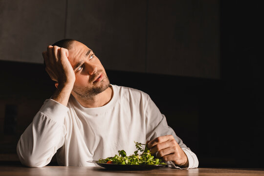 Handsome man sitting at table in kitchen in house. He wear white long sleeve with leaf of salad in his plate. Diet, health food. Man not wanting to eat salad, dream about another food, look at up.