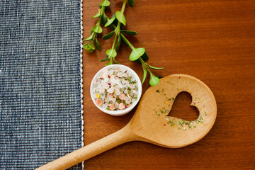 mountain herb salt in a small white container on a dark wooden plate with a heart kitchen spoon on a blue white and pink background, top view, copy space