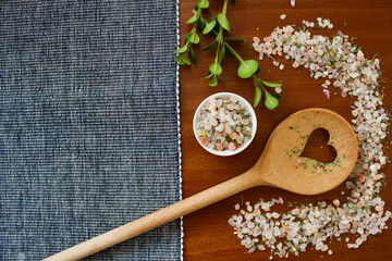 mountain herb salt in a small white container on a dark wooden plate with a heart kitchen spoon on a blue white and pink background, top view, copy space