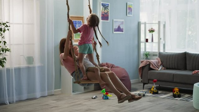 Loving mother sits on swing with son on lap in cozy apartment. Preschooler girl with long braids stands on seat near woman and boy holding ropes