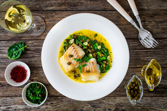 Fish dish - fried cod with spinach and capers in saffron sauce on wooden table
