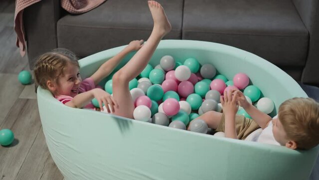 Joyful little children have fun in dry pool with colorful balls in living room. Cute blond brother and elder blonde sister play together at home