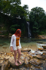 Beautiful girl with red hair enjoying life and beautiful nature. Behind the Abhesi Waterfall, Stones in the mountain river, Kutaisi, Georgia. Vertical photo.