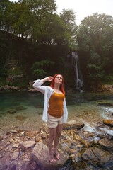 Beautiful girl with red hair enjoying life and beautiful nature. Behind the Abhesi Waterfall, Stones in the mountain river, Kutaisi, Georgia. Vertical photo.