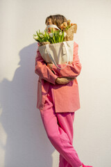 Stylish woman in pink suit carries eco handbag full of flowers on white background. Canvas bag with blank space. Concept of style and spring time