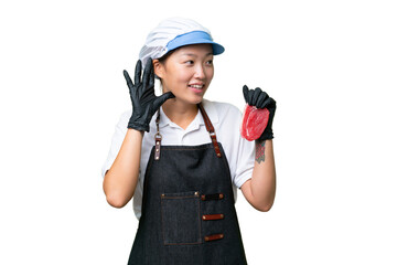 Young caucasian woman wearing an apron and serving fresh cut meat over isolated background listening to something by putting hand on the ear