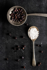 Two old spoons with juniper berries and salt on black background