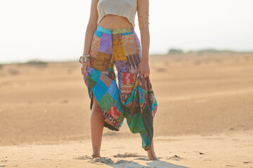 Faceless ethnic woman in colorful outfit on sand. Crop shot of ethnic woman in ornamental outfit and accessories standing on sand in sunlight