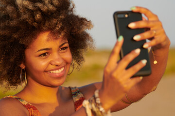 Pretty woman taking selfie via cell phone. Headshot of charming lady with frizzy hair making selfie on mobile phone in sunlight. She is wearing big hoops. Wind blowing her hair.
