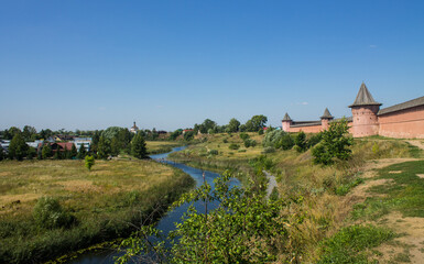 Fototapeta na wymiar the Spaso-Evfimievsky monastery with a brick fortress wall on a hill and the Kamenka river on a green meadow among trees on a clear sunny summer day and a space for copying in Suzdal russia