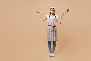 Full body confused shocked young housewife housekeeper chef baker latin woman wear apron toque hat hold in hand laddle spread hand isolated on plain pastel light beige background Cook food concept