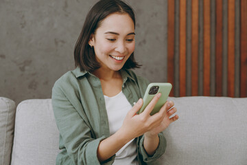 Close up young woman of Asian ethnicity wears casual clothes hold in hand use mobile cell phone sits on grey sofa couch stay at home hotel flat rest relax spend free spare time in living room indoor.