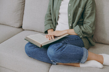 Cropped young smart woman wearing green casual clothes reading book novel sits on grey sofa couch stay at home hotel flat rest relax spend free spare time in living room indoor. Hobby leisure concept.