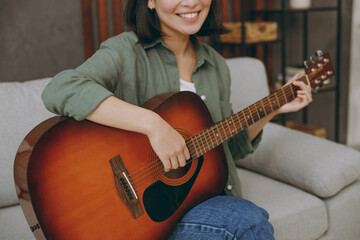 Obraz premium Cropped young smiling happy fun woman of Asian ethnicity wearing casual clothes playing guitar sits on grey sofa couch stay at home hotel flat rest relax spend free spare time in living room indoor.