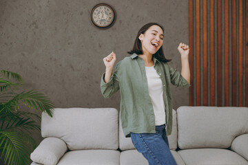 Young smiling fun cool satisfied woman of Asian ethnicity wears casual clothes dancing stand near grey sofa dance couch stay at home hotel flat rest relax spend free spare time in living room indoor.