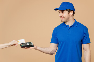 Delivery guy fun employee man wear blue cap t-shirt uniform workwear work as dealer courier hold wireless modern bank payment terminal process acquire credit card isolated on plain beige background.