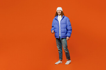 Fototapeta premium Full body smiling happy fun cheerful young man with long curly hair wear hat purple ski padded jacket casual clothes walking going look camera isolated on plain orange red background studio portrait.