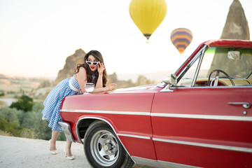beautiful girl in retro style posing near a vintage red cabriolet car on background of balloons in...