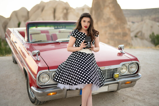 beautiful girl in retro style posing near vintage red cabriolet car with old film camera in hands.